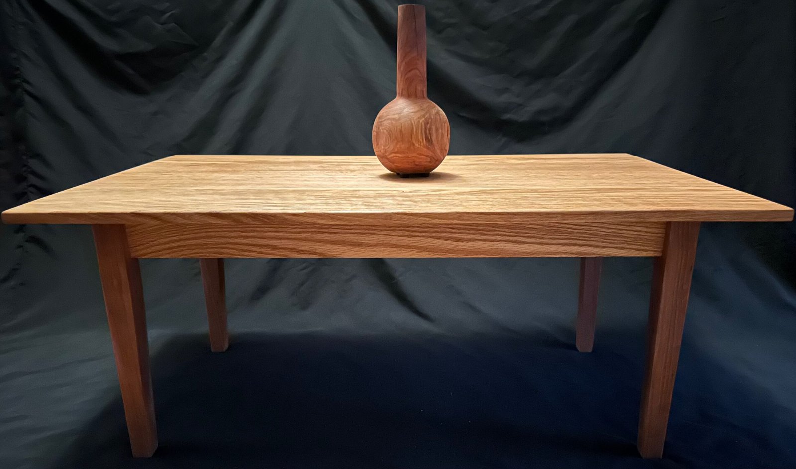 Handcrafted Woodworking, a coffee table with an oak top and tapered cherry legs, with an applewood vase on top. 